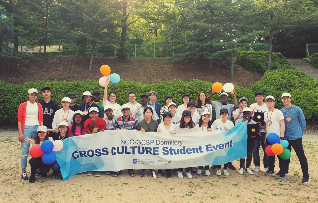 A photo of students (such as Razaul HAQUE) holding event banners and event products at the 2018 dormitory cross culture event held at Jeongbalsan Park